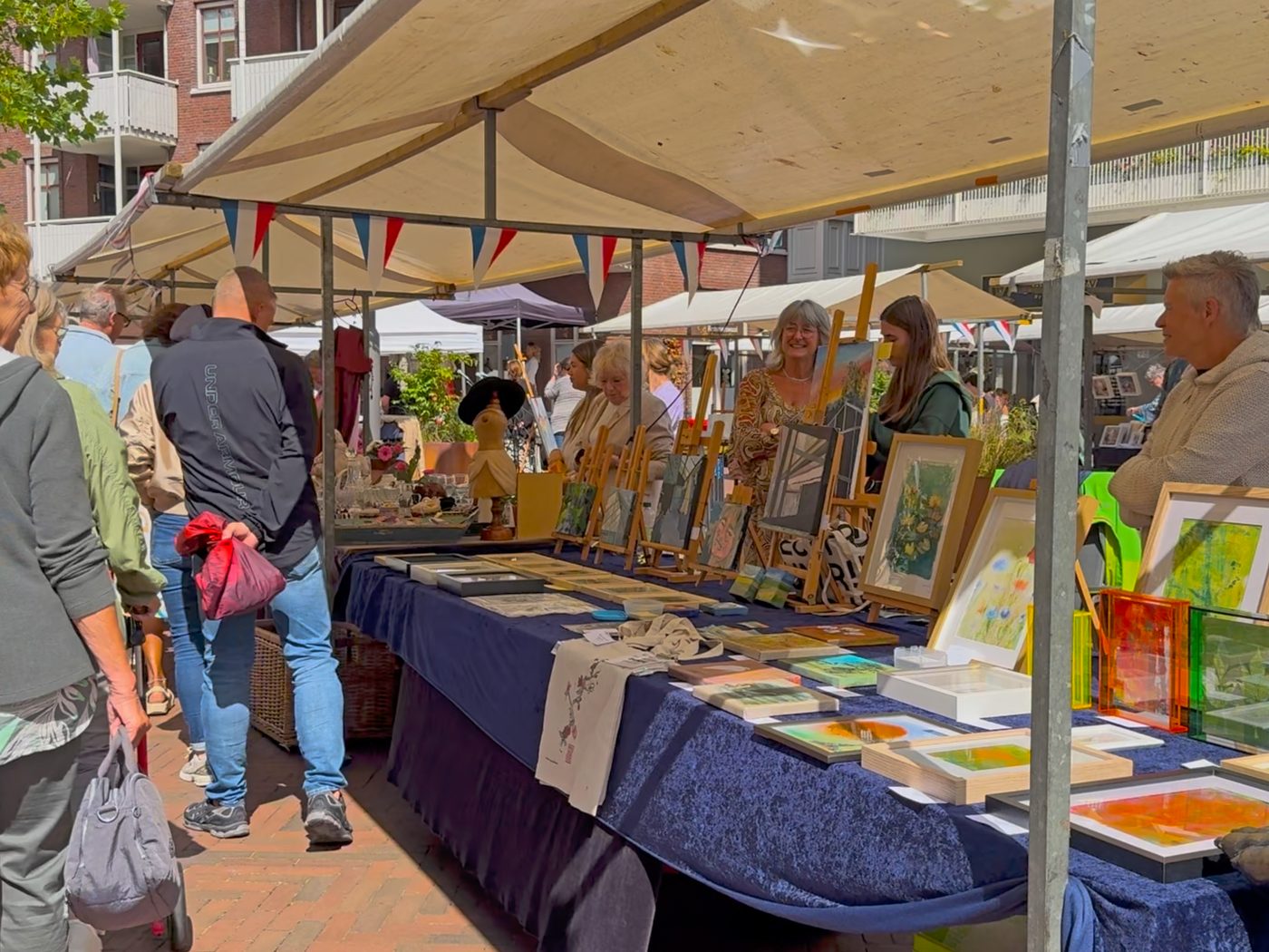 Bezoekers bekijken schilderijen en prenten aan een kunstkraam onder de witte marktkap