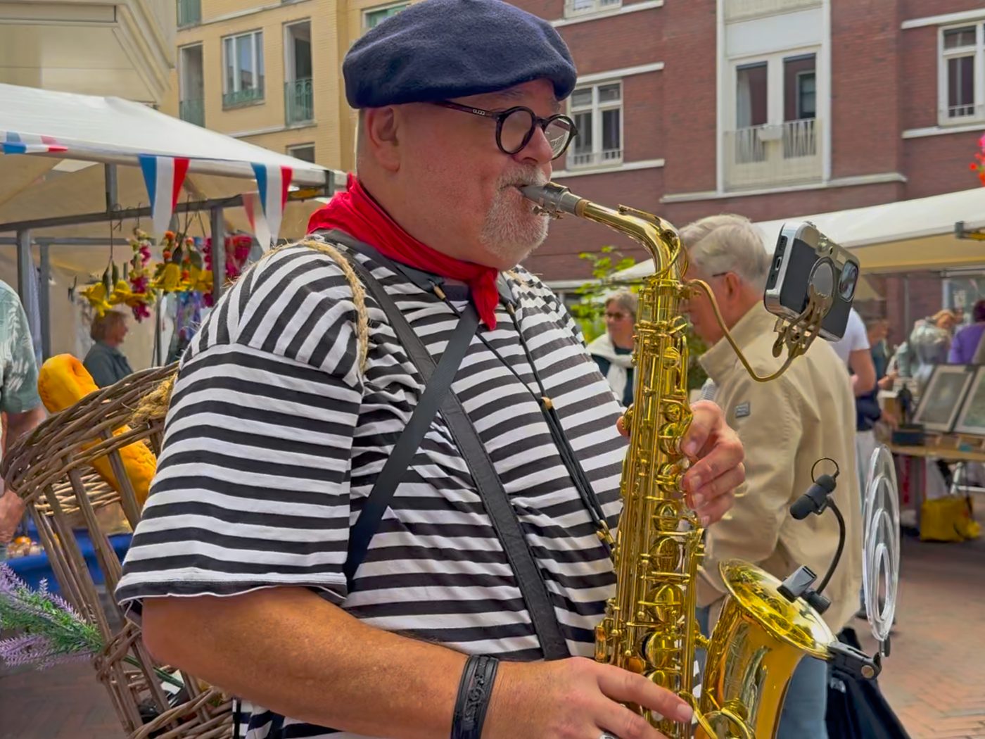 Saxofonist in gestreept shirt met baret en rode sjaal speelt tussen de kramen